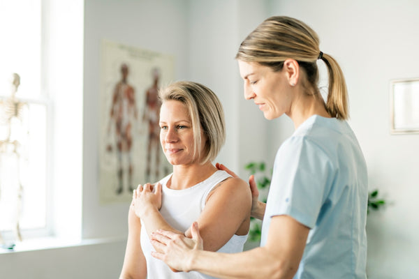 Image of two women. One is a physiotherapist and she is helping the other with her arm and shoulder. The woman receiving physio treatment has her arm across her body to hold the opposite shoulder. They are in a clinical medical setting with anatomical diagrams on the wall behind. 