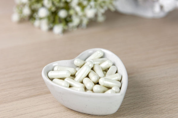 Image of a small white heart shaped ceramic dish containing white coloured capsules of marine collagen. The dish is on a wooden surface and their are some small white flowers in the background. 