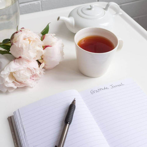 Gratitude journal being written by hand with a black tea and bunch of soft pink flowers