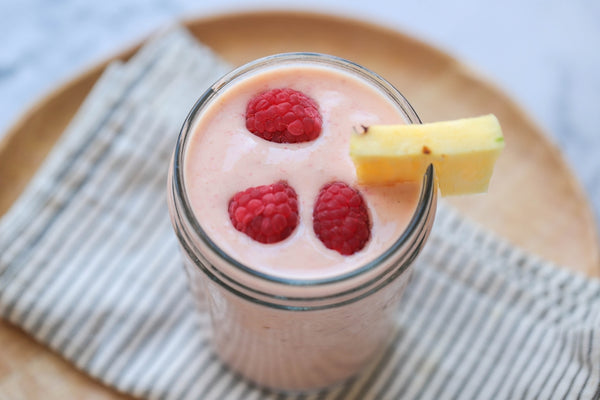 raspberry, mango and pineapple protein smoothie, a pink liquid served in a large glass with 3 red raspberries in the top and a slice of pineapple garnishing the glass