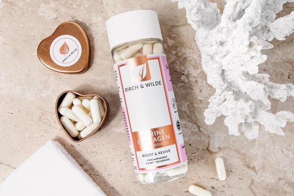 flatlay image from above of a bottle of Birch and Wilde Marine Collagen capsules next to a rose gold heart shaped refill tin with white marine collagen capsules inside and the lid to one side.