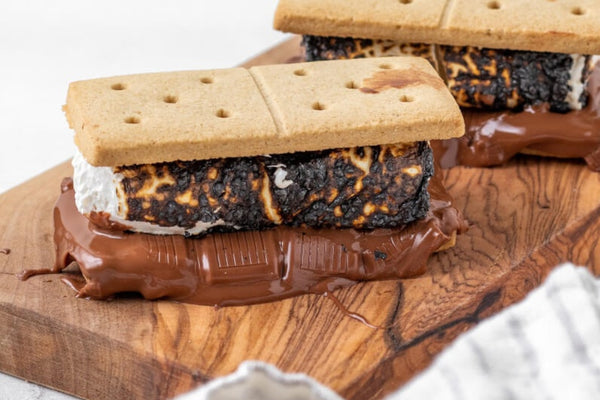 Side view image of a bbq smore with another in the background. Digestive biscuits filled with dark chocolate and marshmallows. Placed on a wooden chopping board with a white background.