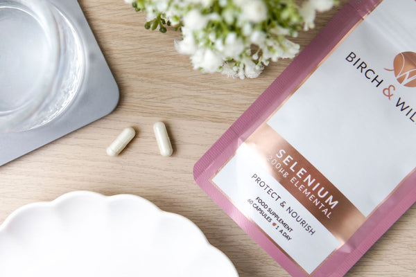 flat lay image of birch and wilde selenium health supplement capsule refill pouch on a wooden background with selenium capsules scattered to the side, and glass of water next to it and some small white flowers