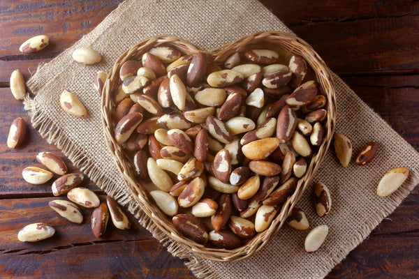 image viewed from above of a large heart shaped basket filled with brazil nuts and other brazil nuts scattered around it. The basket is sitting on a neatly folded kitchen towel and they are both on a wooden work surface