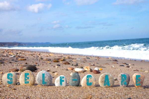 self care painted onto pebbles at a beach with the sea waves sand and blue skies in the background