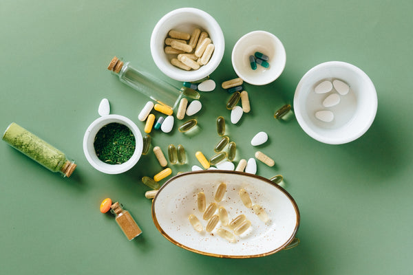 Flatlay image from above of a selection of vitamin and supplement capsules pills and tablets in white pots and scattered loosely on a sage green background