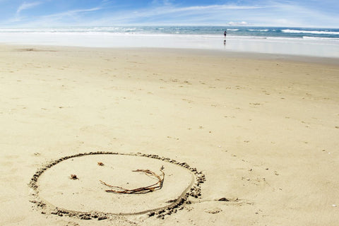 smiling face drawn in flat yellow sand at a big beach with the calm sea in the background to show a happy mood