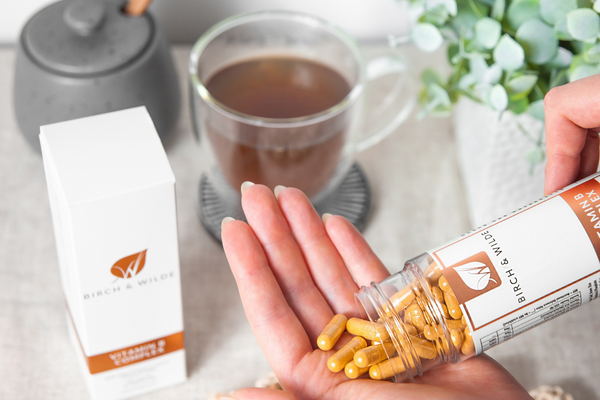 A bottle of birch & wilde vitamin B complex capsules being poured from a clear bottle into and open cupped palm with a cup of tea and a healthy green pot plant in the background