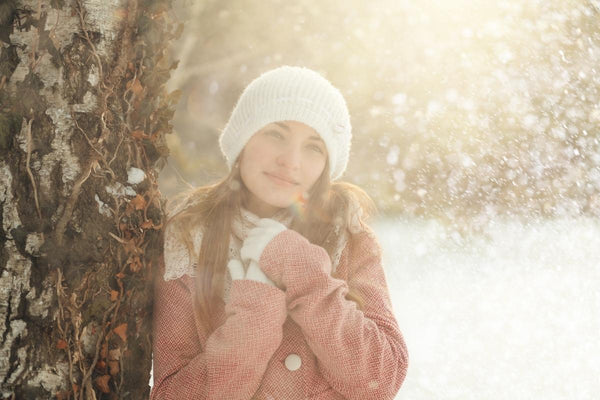 Image of a woman in winter, leaning up against a big tree, wrapped up in a pale pink coat with a white woolly bobble hat and scarf. The sun is hazy through the snow in the background and she has her hands together in front of her to keep warm.