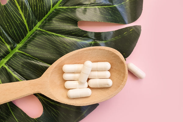 Close up image from above of a large wooden spoon with cream coloured collagen supplement capsules on it and a large green palm leaf behind it, all on a pale pink background