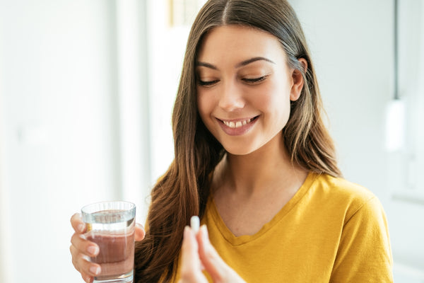 Head shot image of a young women with long straight brown hair holding a glass of water in one hand and a collagen supplement capsule in the other hand whilst smiling and wearing a yellow jumper