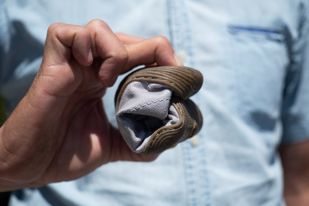 A Wildling minimal shoe rolled up in width is held together by two fingers. In the background the torso of an adult person in a light blue shirt.