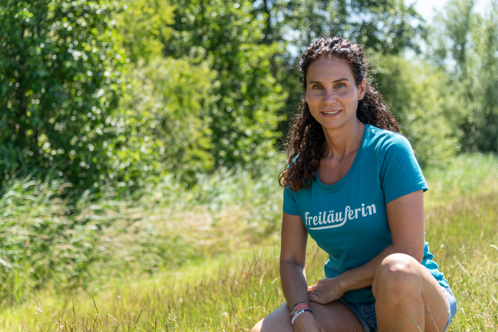 A green meadow, in the background green bushes and trees. On the meadow in squatting position a smiling woman with long dark curly hair and a blue-green t-shirt, which has the inscription 