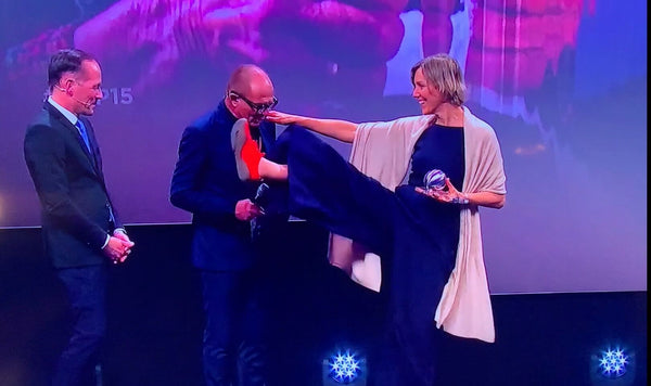 Presenting the German Sustainability Award, Anna Yona on stage with two people in black suits, stretching one leg up to chin level, at her foot a bright red Wildling Shoes minimal shoe.