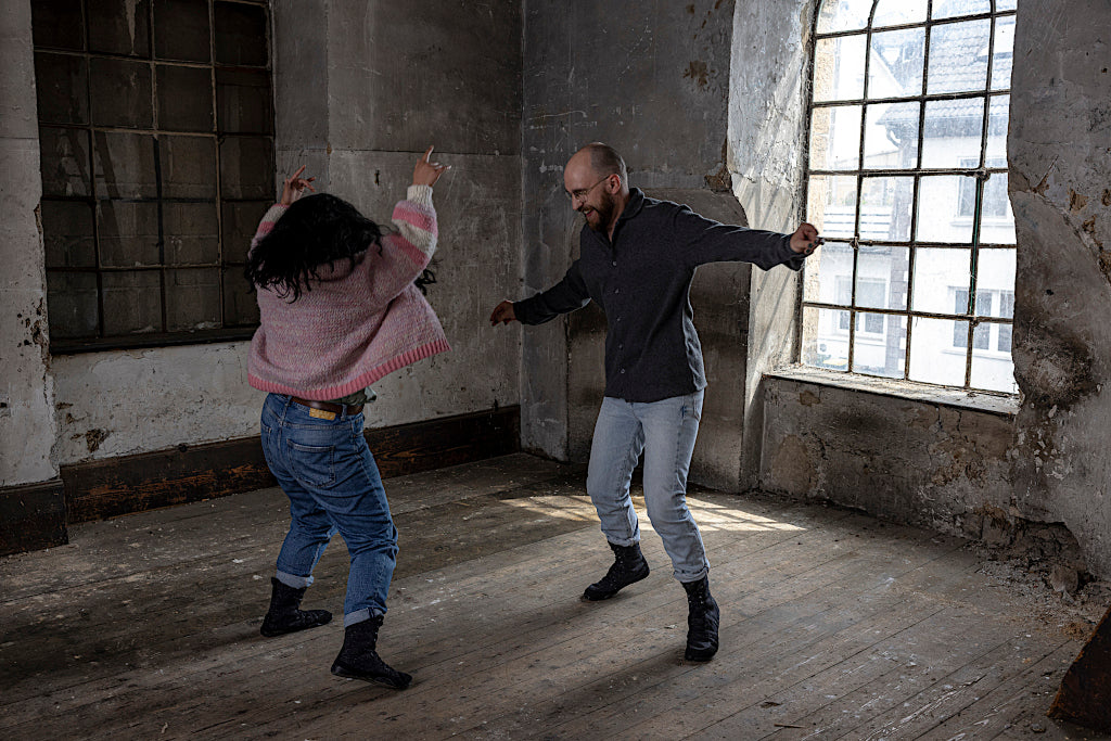 Two people dancing in an empty warehouse in front of a large window on wooden floor.