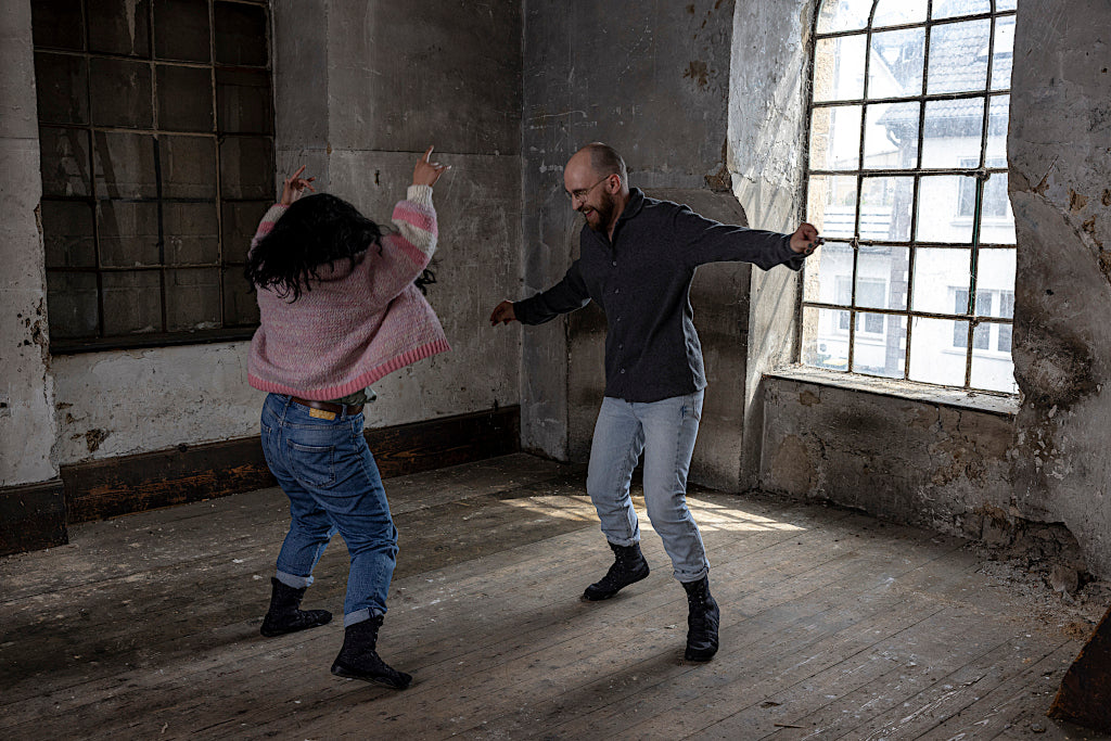 Zwei Personen tanzen in einer leeren Lagerhalle vor einem großen Fenster auf Holzfußboden.