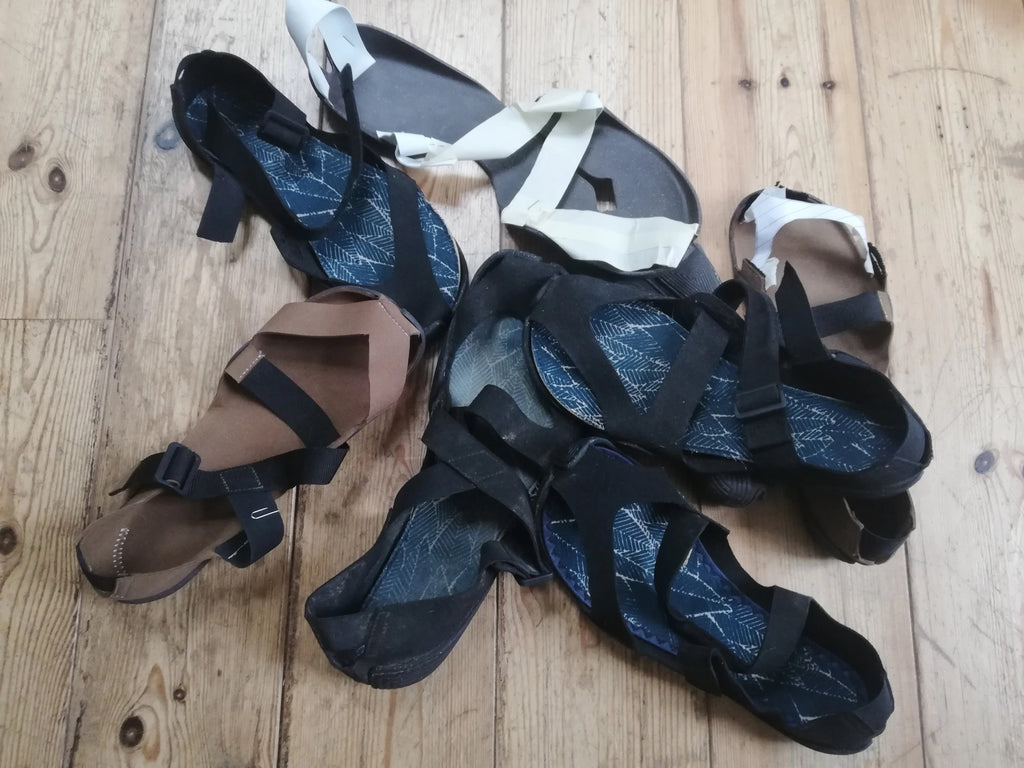 Various Wildling Shoes sandal prototypes lying in a pile on a wooden floor.