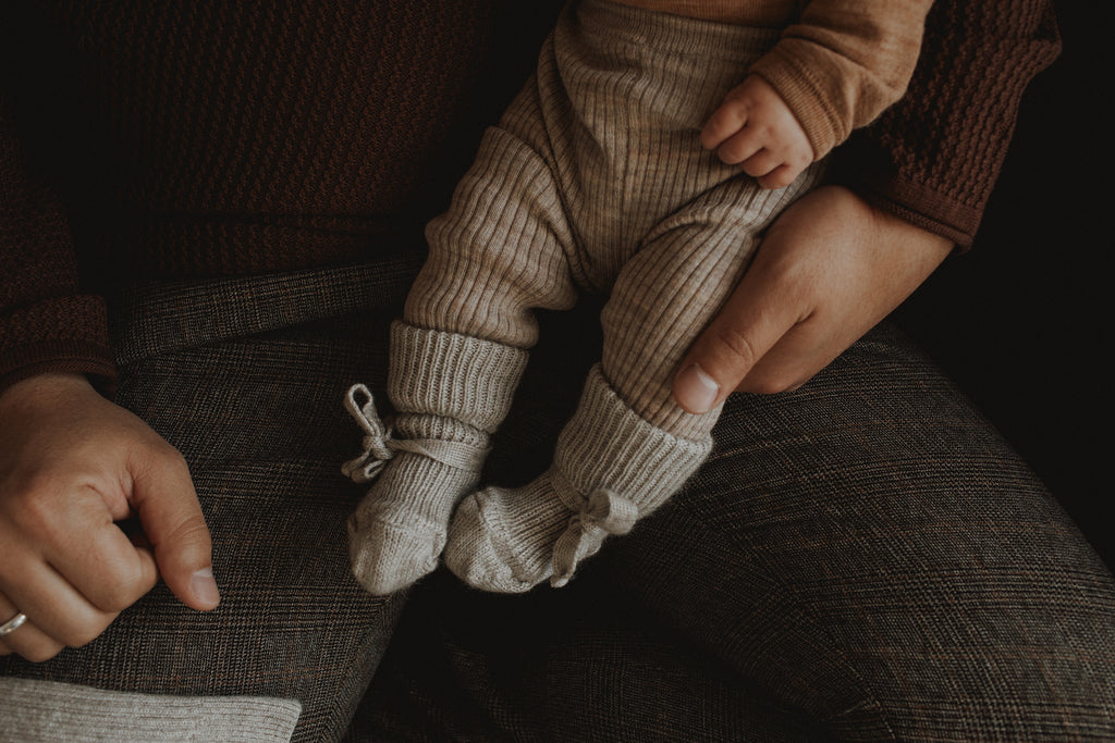 An adult person in cozy, dark brown clothes holds a baby on their lap. The cropped image shows a close-up of the baby's legs wearing cozy brown-gray clothes. The baby's little feet are dressed in light gray knitted baby socks.