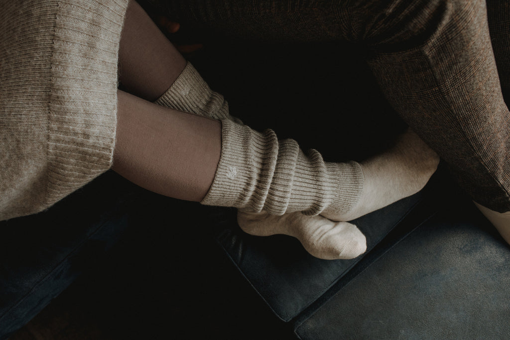A person lies with bent legs on a dark sofa. The cropped image shows the person's legs in close-up. They are wearing light gray knitted woolen cuffs over her socks. Their entire clothing is cozy and warm.