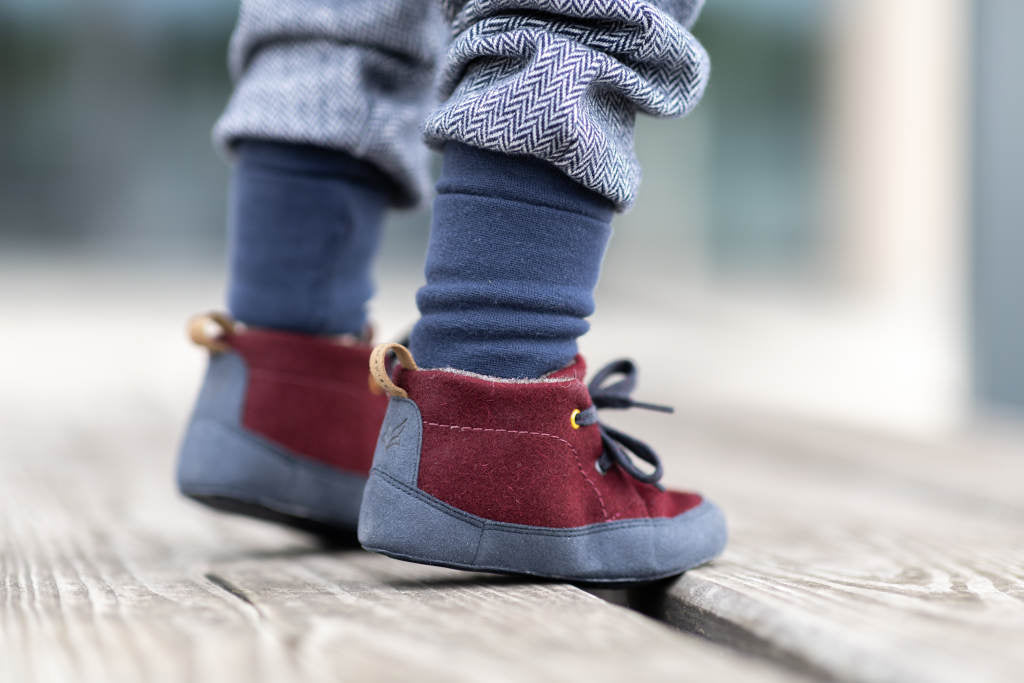 Nahaufnahme von zwei Wildling tragenden Kinderfüßen von hinten. Die Füße stehen auf einem Unterrund aus hellen Holzplanken. Die Beine in blauen Strümpfen und hellblauen Hosen sind etwa bis zu den Waden im Bild.