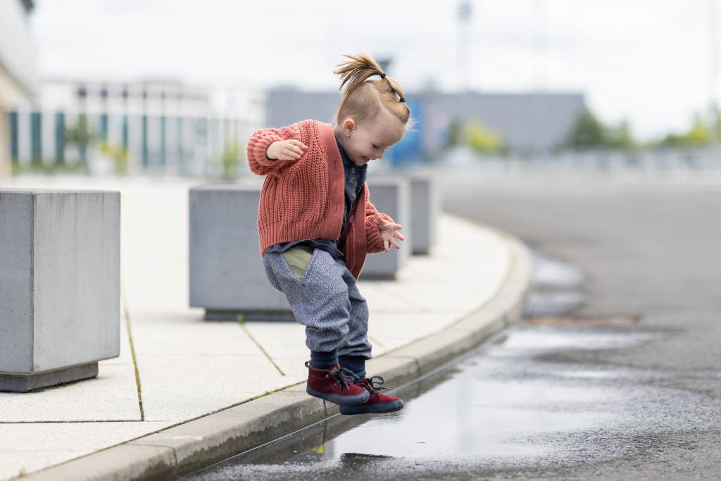 In a bright, urban-looking environment, a child jumps off the curb into a puddle. The child is wearing an ochre cardigan and the Wildling model Douro on their feet.