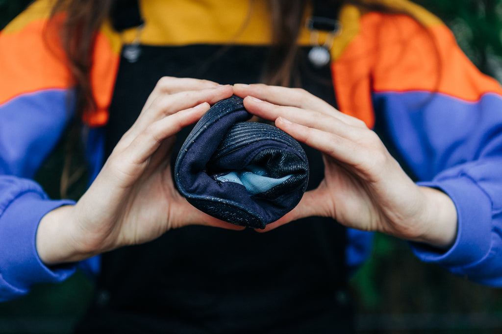 In the foreground there are two hands holding a Renette shoe rolled up like a sushi roll. In the background is the upper body of the person holding the shoe. The person is wearing a sweater with block stripes in yellow, orange and purple from the brand Manitober.