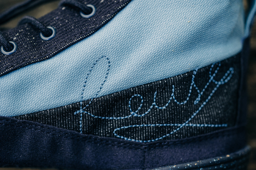 Detail view of the back of the minimal shoe Renette from the side. You can see light blue and dark blue fabric elements and the embroidered word 