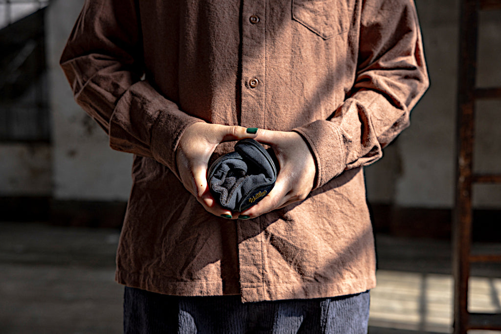 A person in an ocher flannel shirt holds a rolled up Wildling minimal shoe with both hands in front of their chest. In the picture is only the torso of the person, in the background is a dark room, light is coming in from the right.