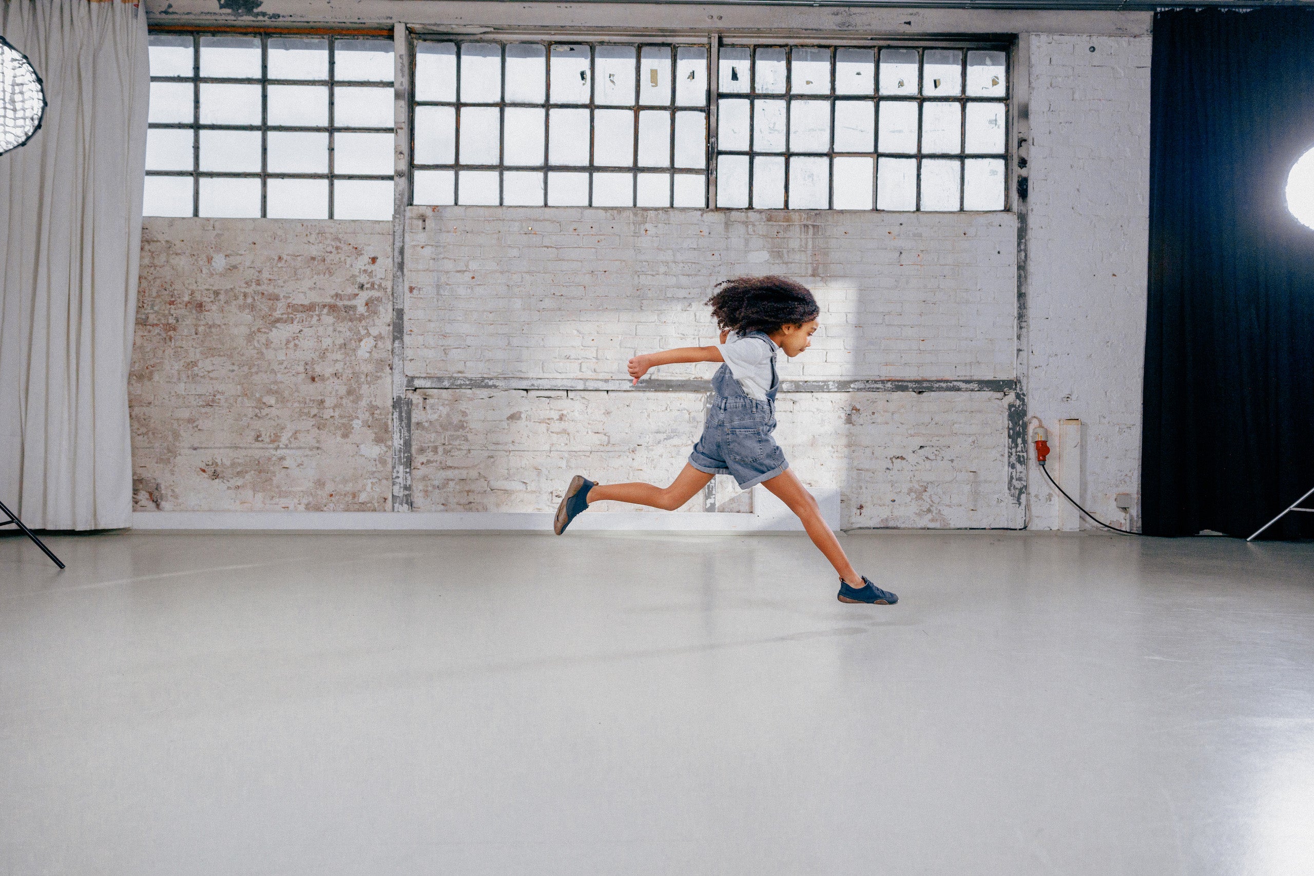 A bright, empty storage room where a child in shorts and Wildling minimal shoes jumps from left to right with his hair flying.