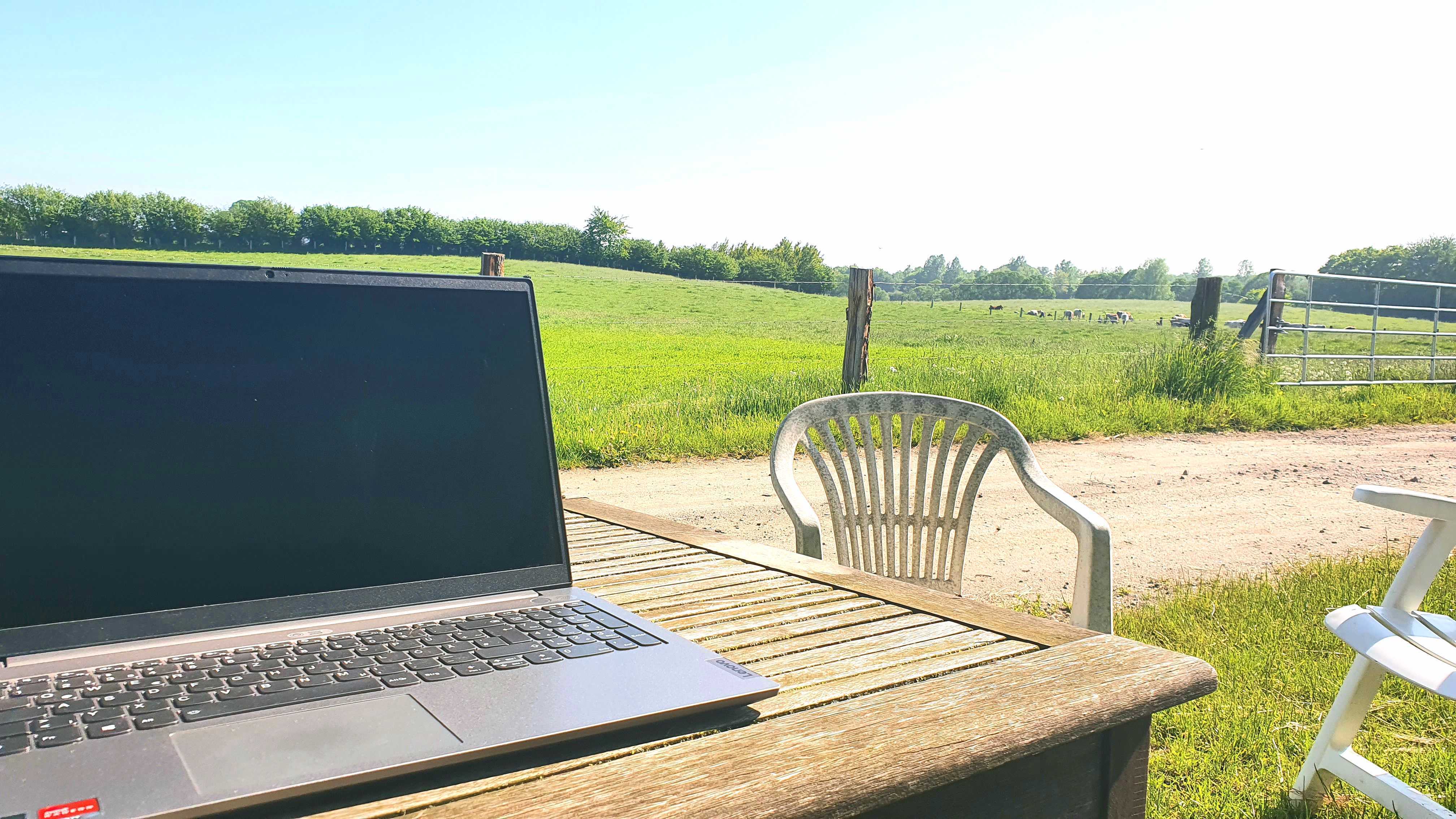 A laptop standing on a wooden table outdoors, behind it meadows and forest view