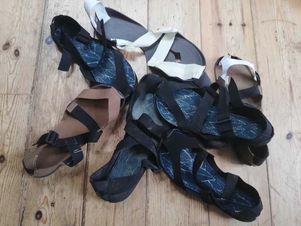 Several prototypes of Wildling minimal shoe sandals on top of each other on a floor of wooden planks.