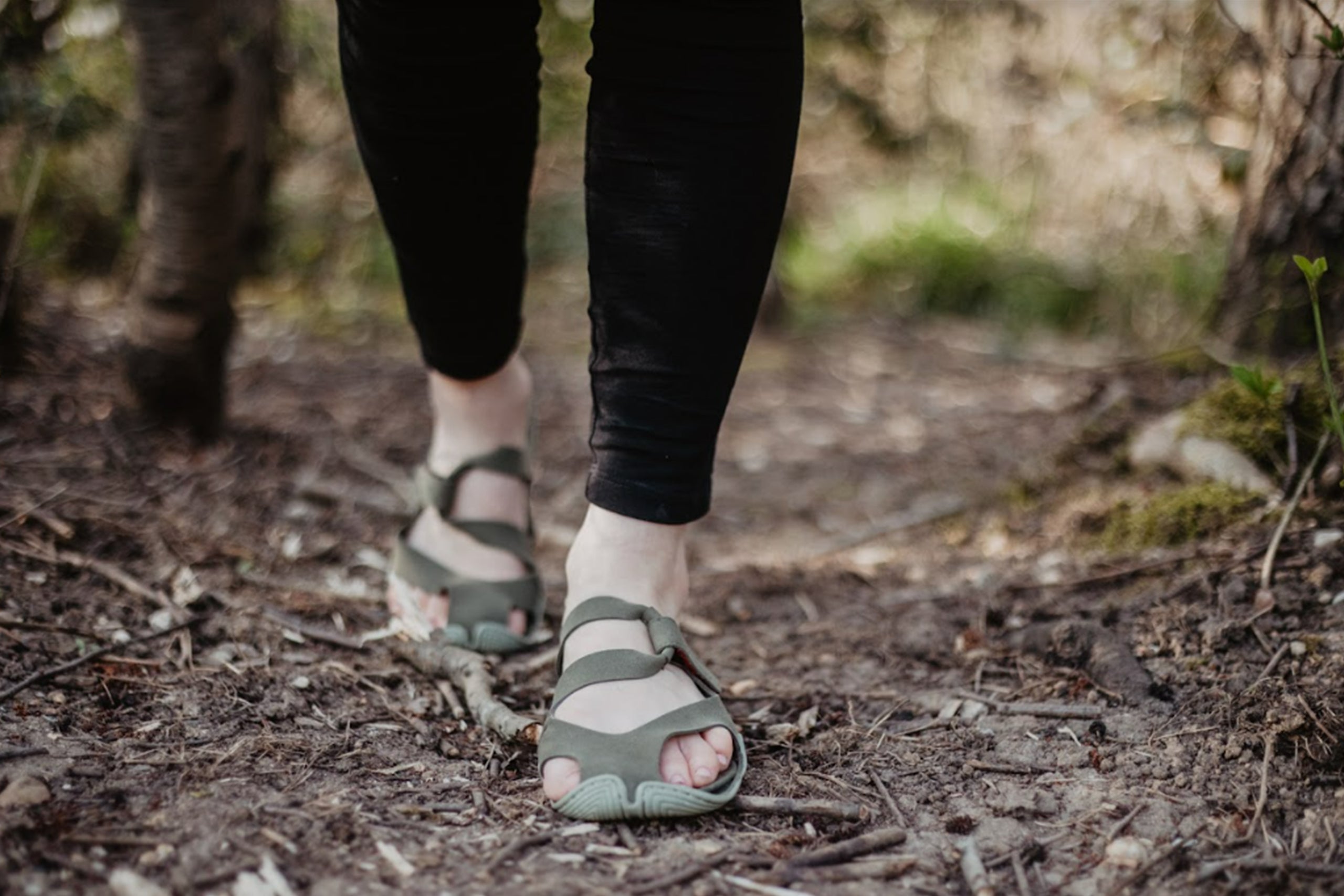 Two feet, about up to the calves in the picture, on a forest path. Wildling sandals on the feet.