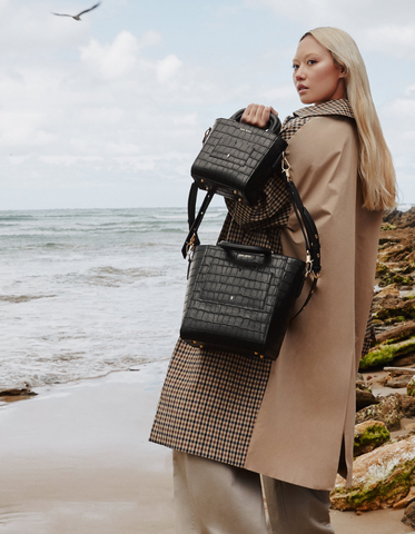 Dechen stands on a beach wearing a beige toned coat and holding two black Sans Beast bags on one arm.
