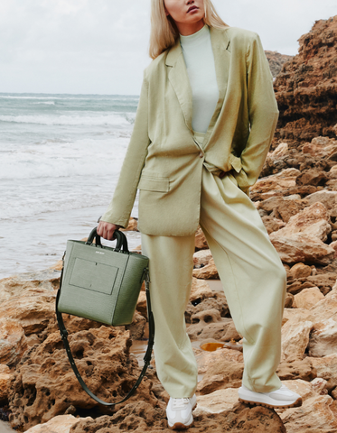 Dechen stands on rocks along a beach, holding a pale green Sans Beast tote bag.