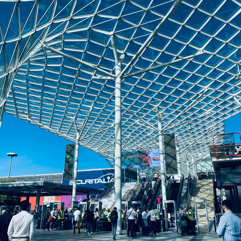 Image of the entrance to Lineapelle trade fair in Milan.  A large metal grid structure stands overhead and a crowd of people walk underneath toward escalators