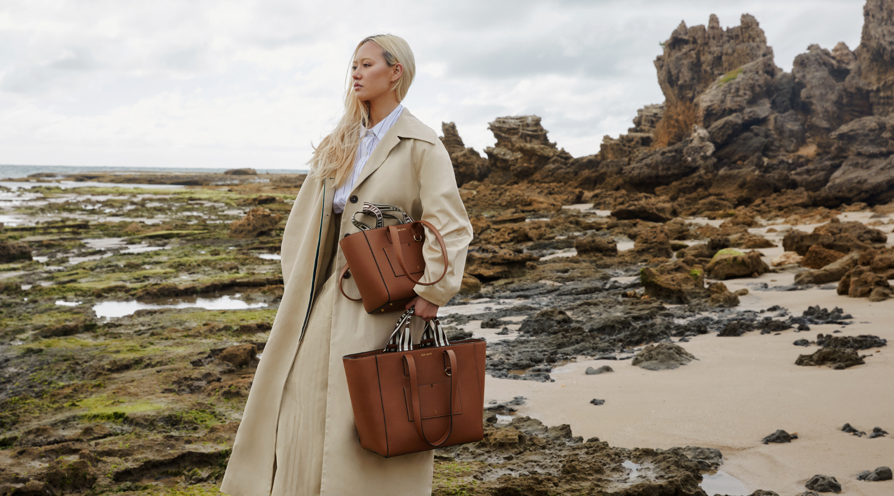 Dechen wears a cream toned trench coat and stands on a rocky beach, holding two brown Sans Beast tote bags