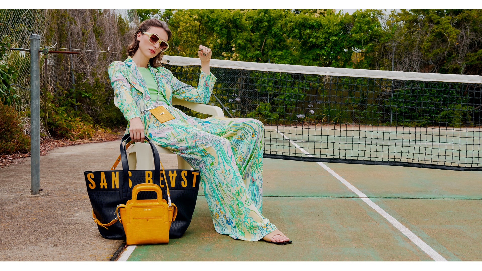 Juliette on a tennis court in a floral pantsuit, with The Activist Tote in Midnight Pebble + The Long Lunch Tote in Mustard Croco.