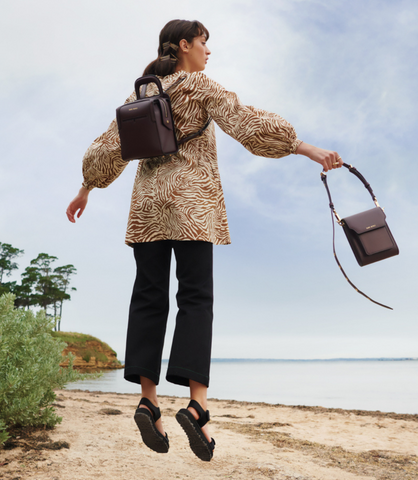 Paris jumps along a beach, holding a burgundy Sans Beast vegan handbag with a matching backpack on her back