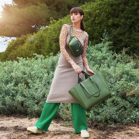 Paris stands on a shoreline wearing a green Sans Beast crossbody bag and carrying a coordinating Sans Beast tote bag