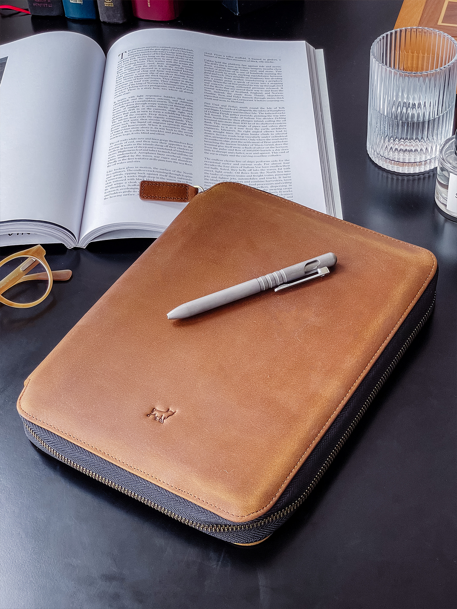 Closed leather notebook with pen on top beside an open book and eyeglasses on a desk.