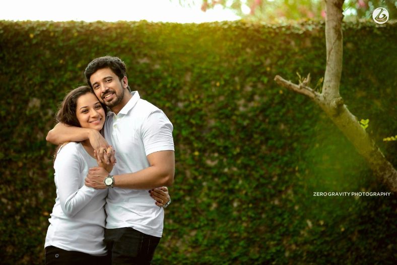 A couple holding red roses poses in beige clothing for an outdoor photo  shoot in a garden surrounded by tall grass by Elyse Zamora in Gilbert, AZ