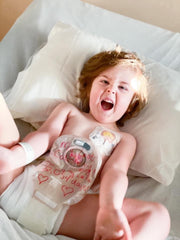Child lays excitedly on hospital bed with colostomy bag exposed.