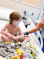 Child looks at nurse as he sits on a hospital bed with blankets around him.