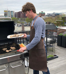 Man grilling chicken on a balcony wearing a leather apron