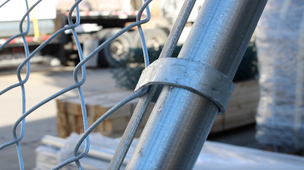 Close-up photograph of a chain link fence post with attached chain link fabric. The fence protects an inventory of wood fence materials.