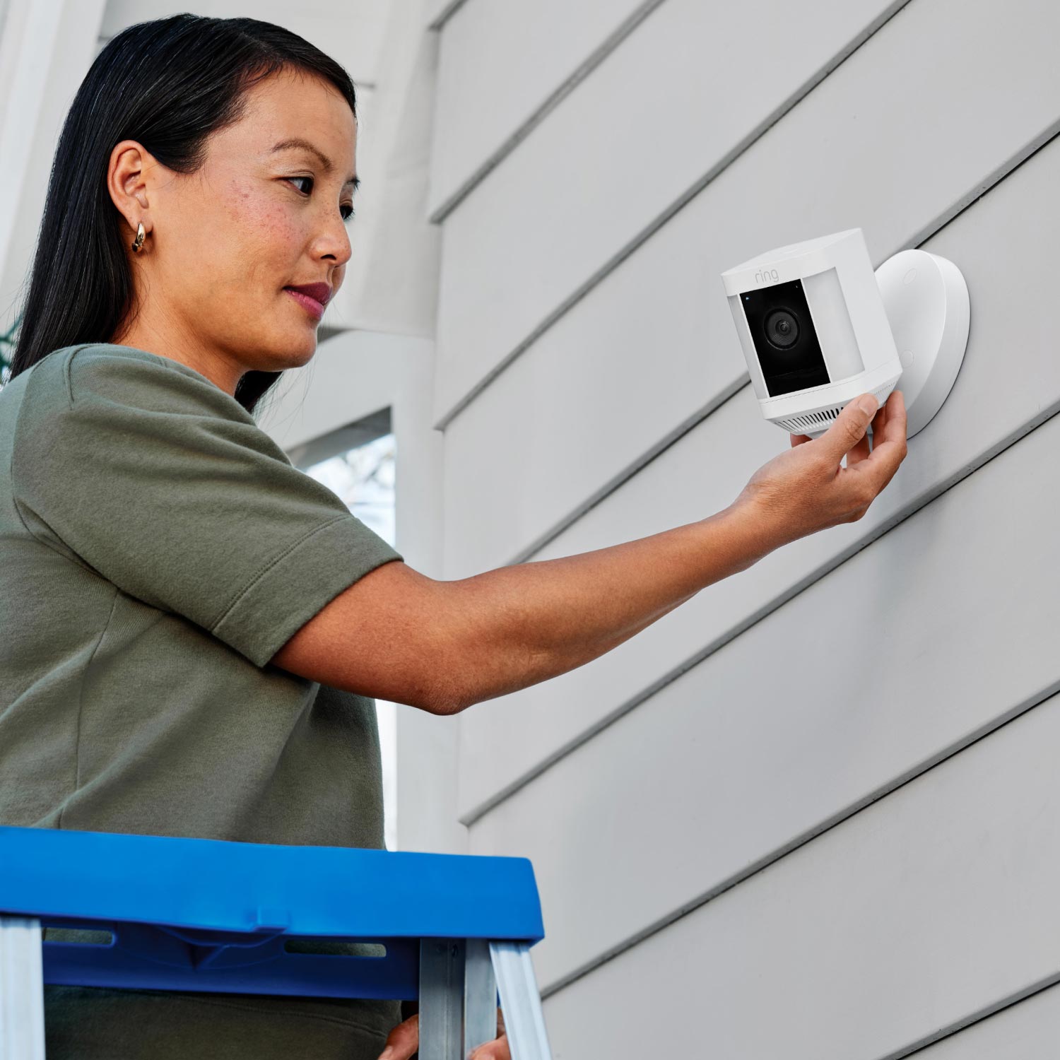 2-Pack Spotlight Cam Plus (Wired) - Woman on a ladder installing Spotlight Cam Plus, Wired model in white on exterior wall of home.