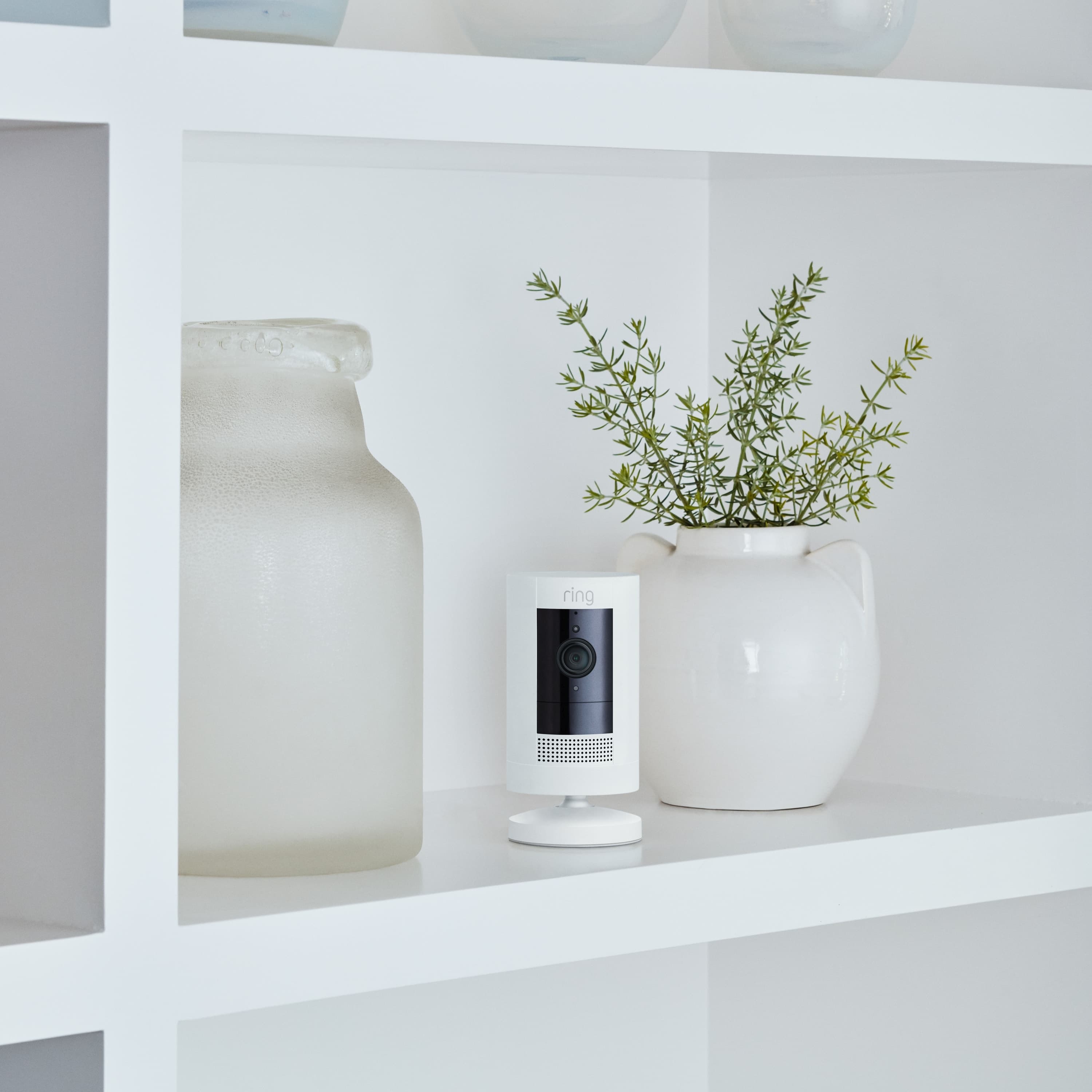 2-Pack Stick Up Cam Battery - Inside a home, Stick Up Cam, Battery model in white, is situated on a shelf between 2 vases.