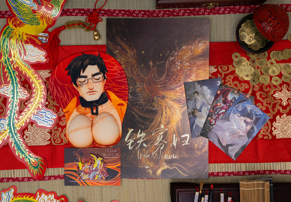 The Shimin 3D mousepad, Phoenix Over Dragon print, Vermillion Bird pin and Iron Trio postcard set on a bamboo mat with an elaborate red table runner surrounded with decorations.
