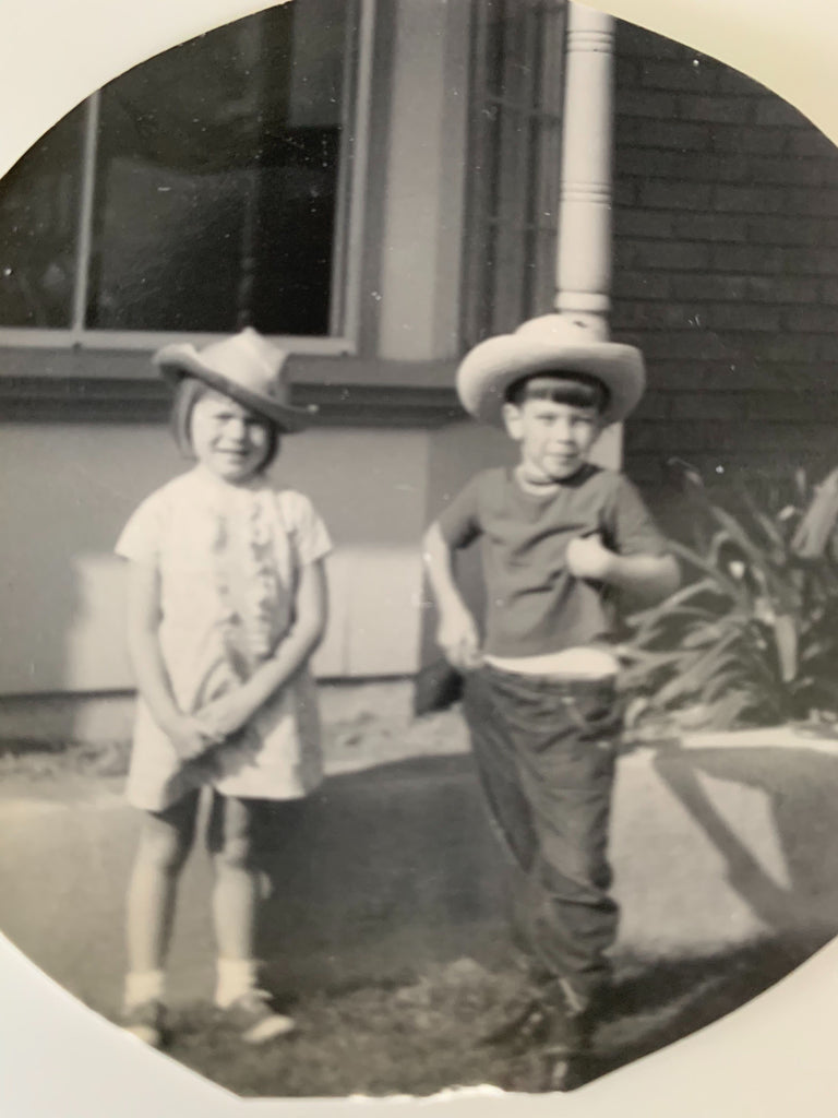 Old black and white photo of little girl and boy wearing cowboy hats next to house.