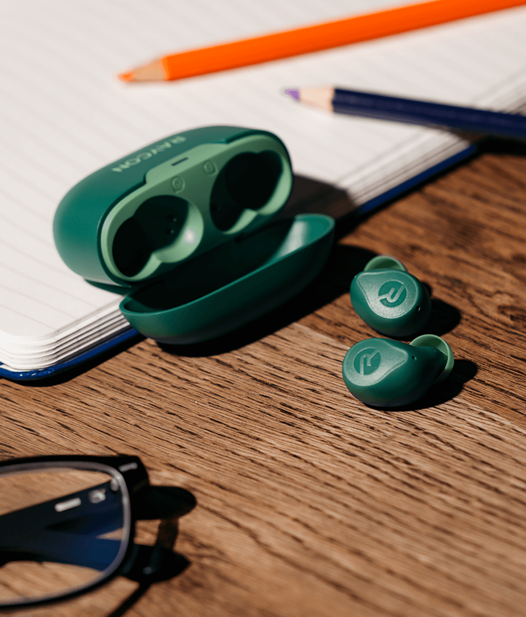 Green wireless earbuds with open charging case on a wooden desk beside glasses and pencils.